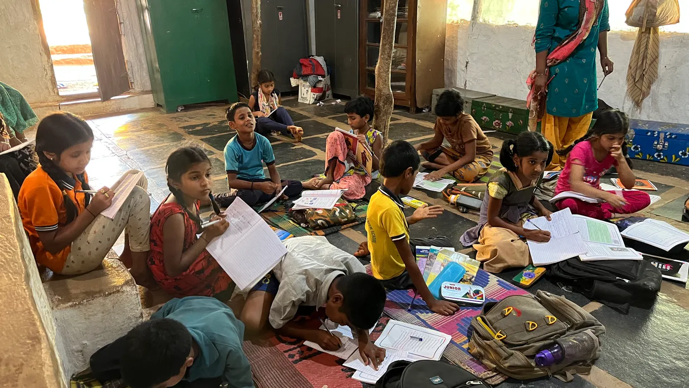 Students sitting on the floor of an Indian classroom writing in notebooks while their teacher watches